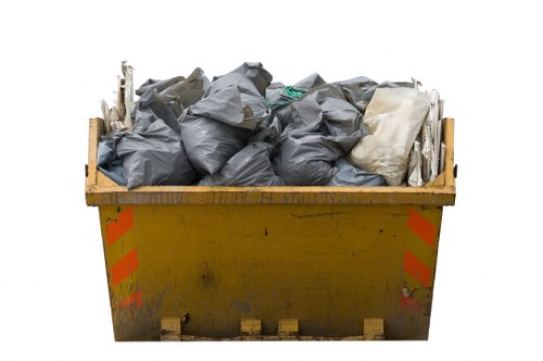 A large yellow skip filled with tightly packed black and grey rubbish bags, some creased and slightly misshapen, with a few white and green items visible among the debris. The skip is positioned outdoors on a paved surface, with orange reflective stripes on its sides indicating it is used for waste collection. The surrounding environment suggests an industrial or commercial area typical of Westminster, with no other structures or objects visible in the background. The overall scene depicts a typical rubbish removal setup managed by Office Clearance Westminster, ready for waste disposal or debris removal services in the local area. The lighting appears natural, with no shadows or reflections indicating overcast weather or diffuse daylight, emphasizing the straightforward and functional appearance of the waste container.