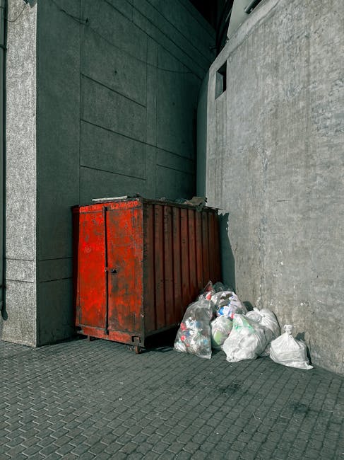 A small, burgundy flatbed truck parked on a city street in front of a modern office building with large glass windows. The truck's open cargo bed is filled with various types of waste materials, inclu