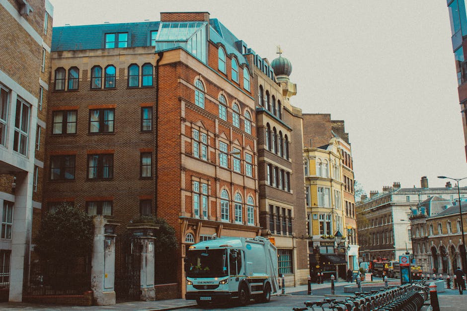 A city street scene featuring a light blue street cleaning vehicle parked alongside historic multi-storey buildings with brick facades and ornate windows. The vehicles are situated on a paved road bor
